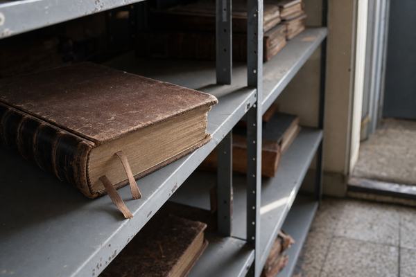 Digital press photograph taken in 2014 inside a county records vault, full-frame DSLR with 50mm lens at f/2.2, ISO 1600 showing slight luminance noise. Asymmetric composition with the dominant subject placed on the left third: a worn, leather-bound registry ledger resting on a painted steel shelf, ribbon markers protruding. Raking afternoon light from a high window picks up dust motes and scuffs in the gray paint; out-of-focus ledgers recede into shadow. Incidental background details: chipped concrete floor edge and a steel doorframe just visible to the right, no signage. Shallow depth of field, natural white balance; no text or lettering visible anywhere, no watermarks.