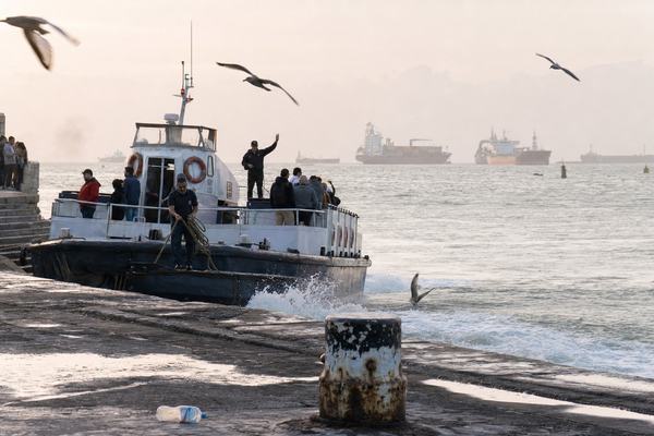 Digital press photograph (2025) at dawn on the ferry steps along Calle de la Barquillera in Cádiz; a worker-owned Bay ferry, the San Isidro, easing away from the wet stone steps under pale autumn light; the captain on deck counting passengers with one hand raised while a crewman coils a line; gulls cutting across the frame; in the distant haze beyond the bay mouth several large container ships and a tanker sit idle at anchor; oblique composition from the steps with a foreground bollard showing chipped paint and rust streaks, puddles reflecting the sky, a stray plastic bottle near the curb; shot on a full-frame mirrorless with a 35mm lens, ISO 3200, 1/250s; slight motion blur in water and birds, mild high-ISO noise in shadows, gentle backlight flare on spray; candid, unposed; no legible text, no signage, no watermarks.