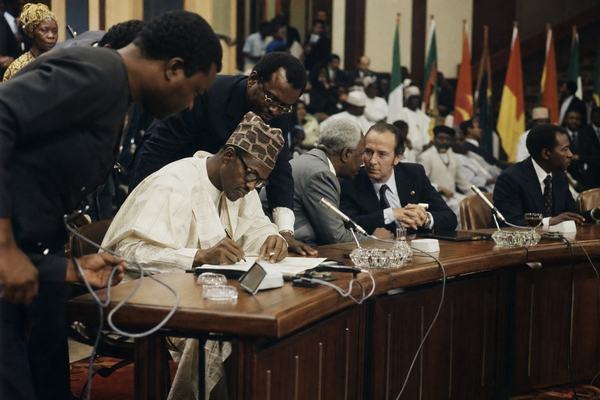 Archival color photograph (1975) shot on Ektachrome 100 with a Nikon F2 and 35mm lens, warm tungsten cast and moderate grain, slight edge vignetting and a touch of motion blur in hands. Asymmetric composition from the left aisle inside the Lagos National Theatre hall: a West African head of state signs at a long table while another leader leans in to read, an elder statesman murmurs to a foreign minister just behind; aides hover with cables and unlabelled desk microphones visible. Background flags without any readable emblems, scuffed parquet floor, scattered glasses and ashtrays, spectators blurred in the rear. No text or signage visible; distinct faces and natural posture; indoor ambient light only.