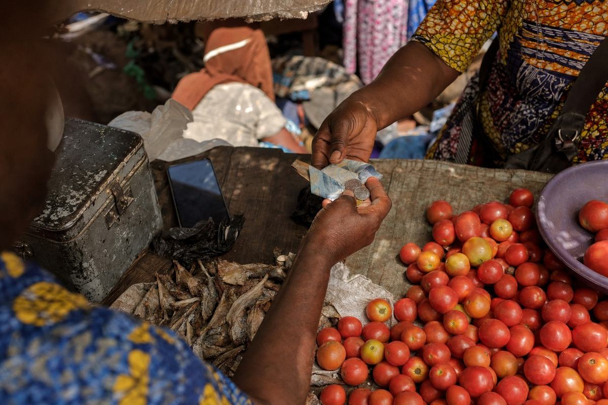 Digital press photograph (2025) with a 35mm prime lens at f/2, shallow depth of field and light high-ISO noise. Oblique top-down angle at a cross-border market stall on the Abidjan–Lagos corridor: two traders’ hands exchange folded ECO banknotes and a few coins; the notes are angled and folded so no lettering or numerals are visible. A small dented steel cash box with scratches sits beside heaps of tomatoes and dried fish; a phone lies face-down with the screen off. Weathered tarpaulin shade overhead, dust motes visible, shoppers and hanging fabric patterns blurred in the background. No visible text or logos anywhere; one dominant action is the handover of money.