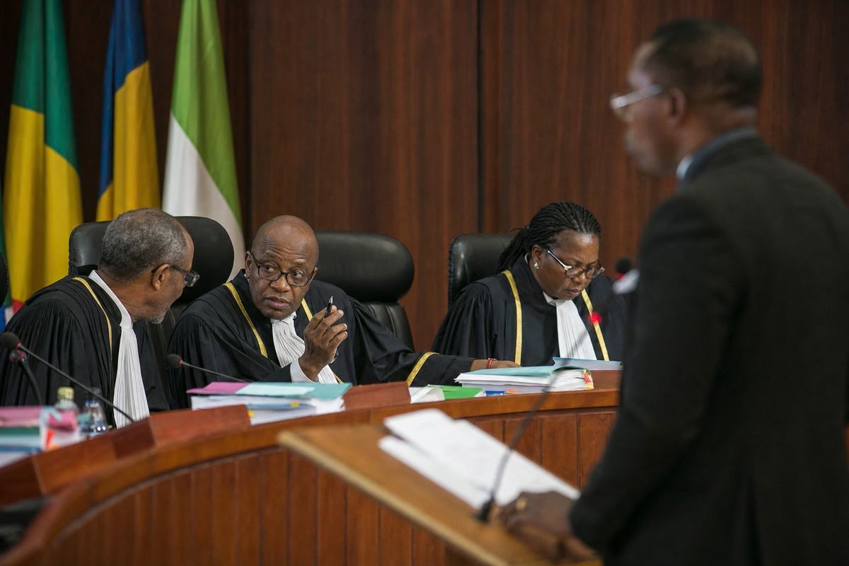 Digital press photograph (2019) with an 85mm lens, shallow depth of field and natural window light. Interior of the West African Court of Justice: three judges in robes on a curved bench, the presiding judge mid-gesture with a pen while a colleague leans to confer; counsel stands at a lectern in profile. Case files stacked with colored tabs, microphones, and wood paneling; small flags behind the bench turned so no emblems or lettering are readable. Papers on the lectern are out of focus, no visible text anywhere; eyeglass glare and natural skin texture evident.