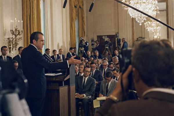 Archival color photograph, White House East Room, June 1971; candid off-center composition with President Richard Nixon at a wooden lectern mid-gesture on the left third of frame; network television cameras, boom mics, and a photographer’s shoulder partially obscuring the foreground; rows of reporters in period-correct suits and horn-rim glasses taking notes; Secret Service agents spaced along gilded walls; chandeliers producing slight halation; 35mm Ektachrome 100 film with warm tungsten cast, visible grain, and minor motion blur in a reporter’s hand; shot from press pool height with a 50mm lens, slight tilt; uneven exposure near tall windows; no legible text or logos.