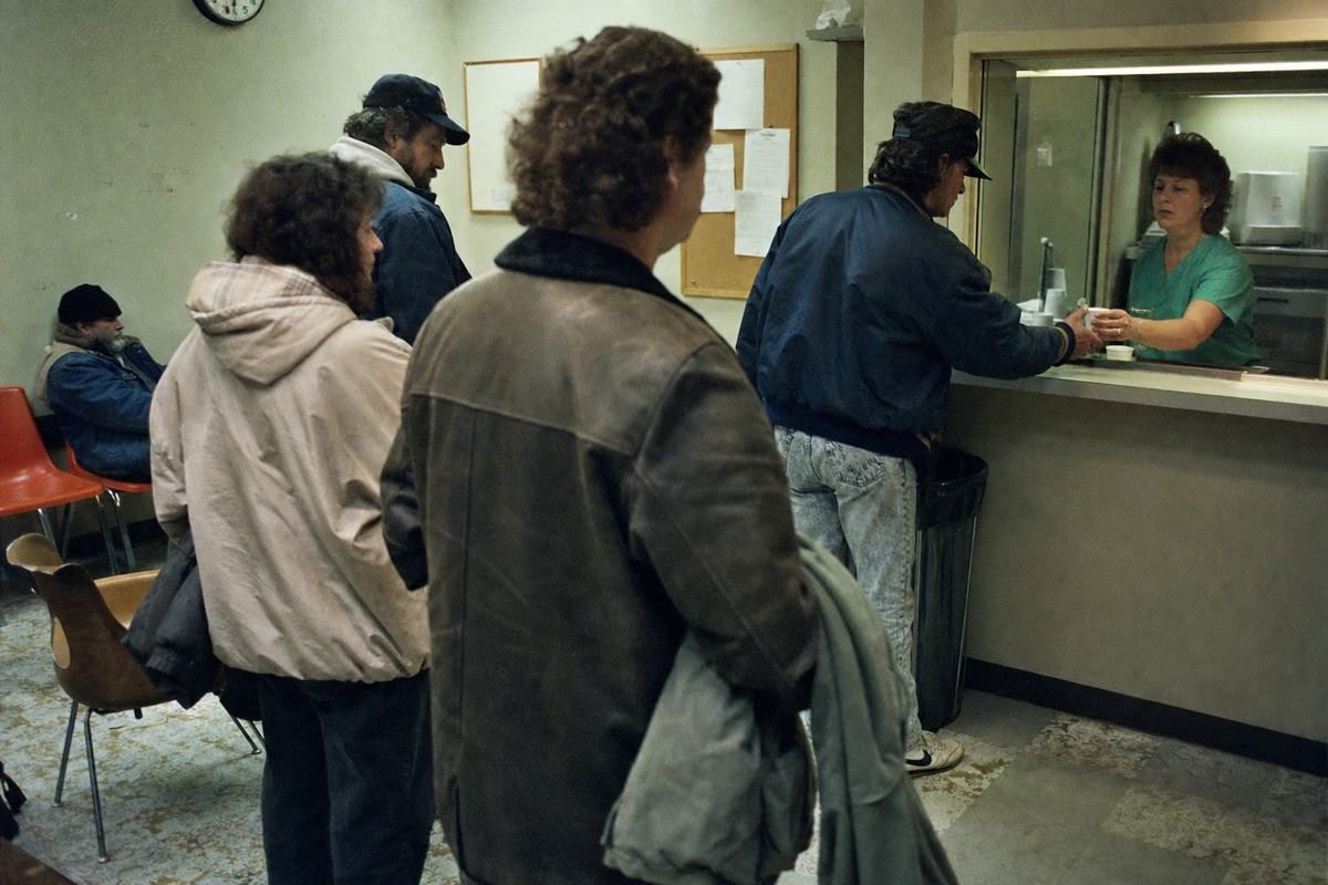 Color documentary photograph inside a community addiction health center, circa 1986; asymmetrical framing of a methadone dosing window at right, nurse in scrubs handing a small paper cup through plexiglass; a short line of patients of varied ages and builds, winter coats draped over arms; scuffed linoleum floors, mismatched plastic chairs, a corkboard with pinned papers (no legible text), and a wall clock partly cropped; fluorescent lighting with green cast; 35mm Fujicolor 400 film with pronounced grain and slight motion blur; shot at 35mm focal length from shoulder height; no logos or readable signage.