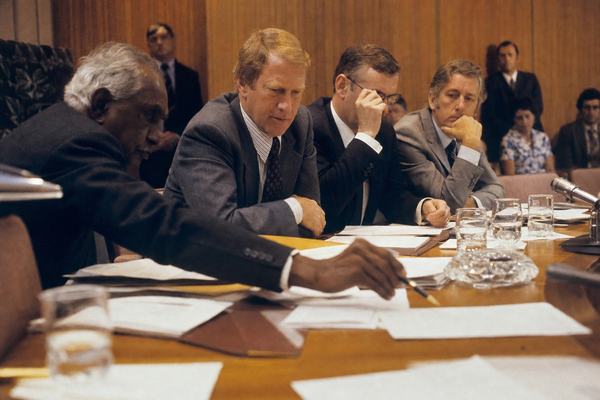 A 1972 color photograph inside the provisional Treaty Chamber at Old Parliament House, Canberra, captured on 35mm Kodachrome 64 with a Nikon F2 and 50mm lens from a low oblique angle along the table. Asymmetric composition: foreground left shows an elder’s hand reaching with a fountain pen toward treaty papers; mid‑frame three ministers lean in, one adjusting his glasses; background has wood‑panelled walls and a few quiet onlookers slightly out of focus. Warm tungsten cast, fine Kodachrome grain, slight motion blur on hands, shallow depth of field isolating the gesture. Water glasses, leather folders, and an ashtray catch the light. No signage or readable text on documents visible.