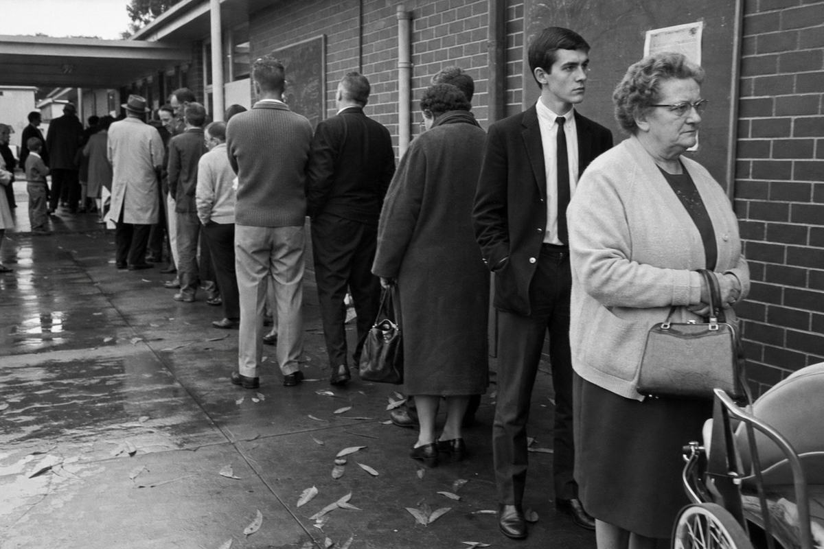 A 1967 black‑and‑white press photograph outside a suburban Melbourne primary school used as a polling place on 27 May 1967. Shot on 35mm Kodak Tri‑X 400 pushed to 800 with a Leica M3 and 35mm lens. Asymmetric composition: a queue bends along a damp brick wall; foreground right shows an elderly woman in a cardigan clutching a small handbag, a young man in a narrow tie behind her. Overcast winter light, wet pavement with scattered eucalyptus leaves, a pram wheel just entering frame. Posters and a chalkboard are partially visible but turned away and unreadable. Moderate grain, edge vignetting, slight motion blur as the line shuffles forward.