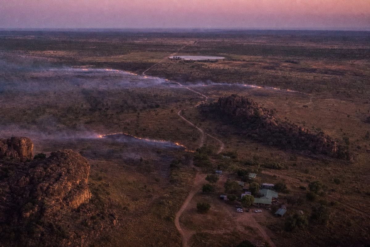 A 2016 digital aerial photograph at dusk over a jointly managed national park in the Northern Territory, shot from a helicopter with a Canon 5D Mark III and 70–200mm lens at 1/500s, ISO 3200. Asymmetric composition: ranger station buildings and two parked utes cluster at lower right; thin, glowing lines of cool burns trace across open savannah; sandstone outcrops cast long shadows; a dirt track winds to a distant billabong under mauve sky. Fine high‑ISO luminance noise, slight motion blur at frame edges from aircraft vibration. No signage or lettering visible.