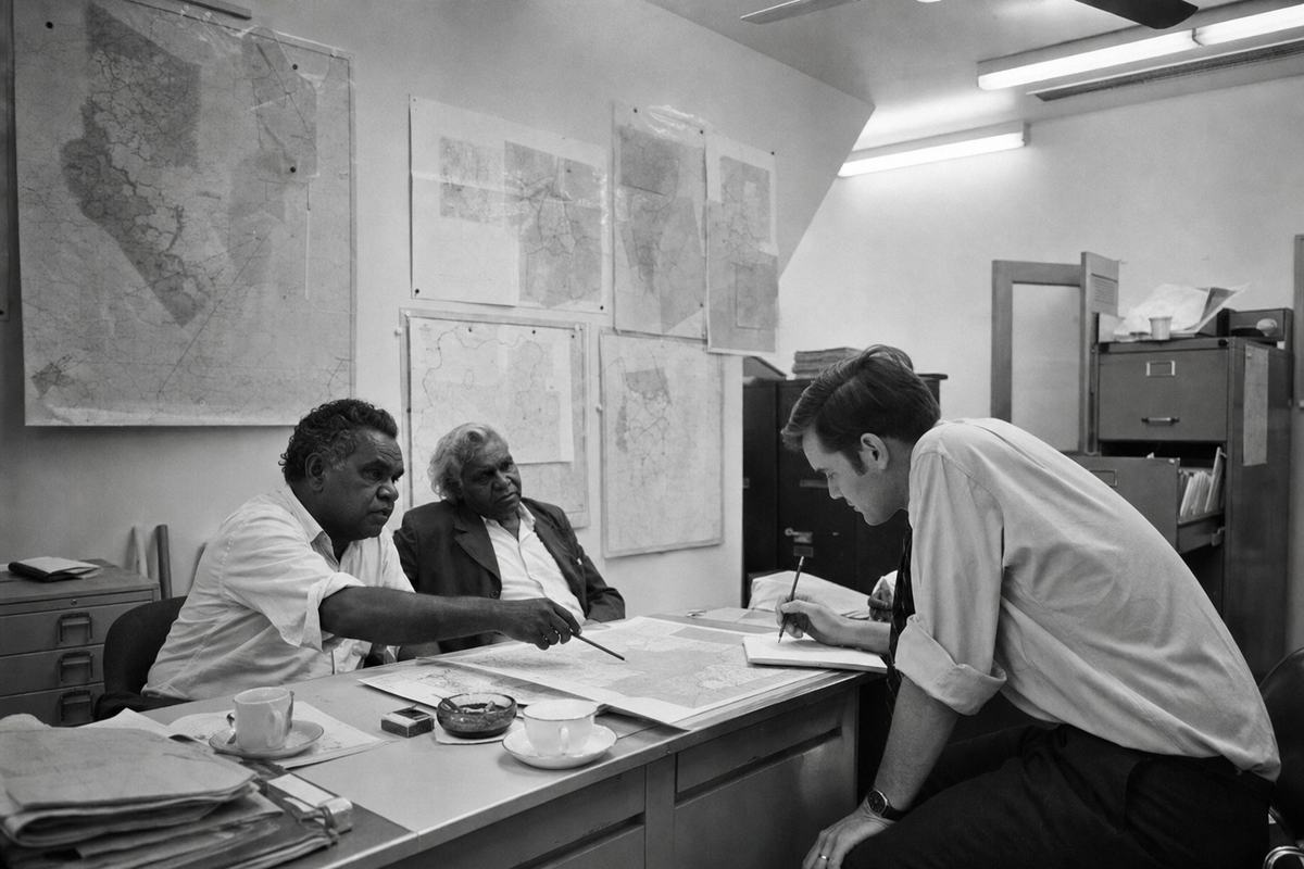 A late‑1960s black‑and‑white documentary photograph inside the National Treaty Commission office in Canberra. Shot on 120 Ilford HP5 with a Rolleiflex 80mm lens, soft contrast and fine grain. Asymmetric composition: two visiting elders sit at a metal desk on the left; one gestures with a pencil to a paper map while the other leans back slightly. A young public servant on the right bends over, listening, pencil poised above a notepad. Walls are crowded with pinned maps and acetate overlays; a ceiling fan and fluorescent tubes overhead; open filing cabinet drawers, teacups, and an ashtray on the desk. No readable text on maps or folders.