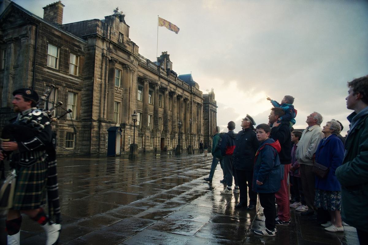 35mm colour photograph on Fujicolor Superia 200, April 1996; oblique street-level view of Parliament House, Edinburgh, wet flagstones reflecting a break in overcast light; the royal standard flies from the roofline; a small crowd at the right edge—students in jackets, an elderly couple, a uniformed constable—faces distinct; a child on a parent’s shoulders points up toward the flag; a piper in a muted kilt strides out of frame left; 35mm lens with slight barrel distortion; fine grain, natural saturation with a faint green cast in shadows; minor handheld motion blur in two passersby; no banners, plaques, or text visible.
