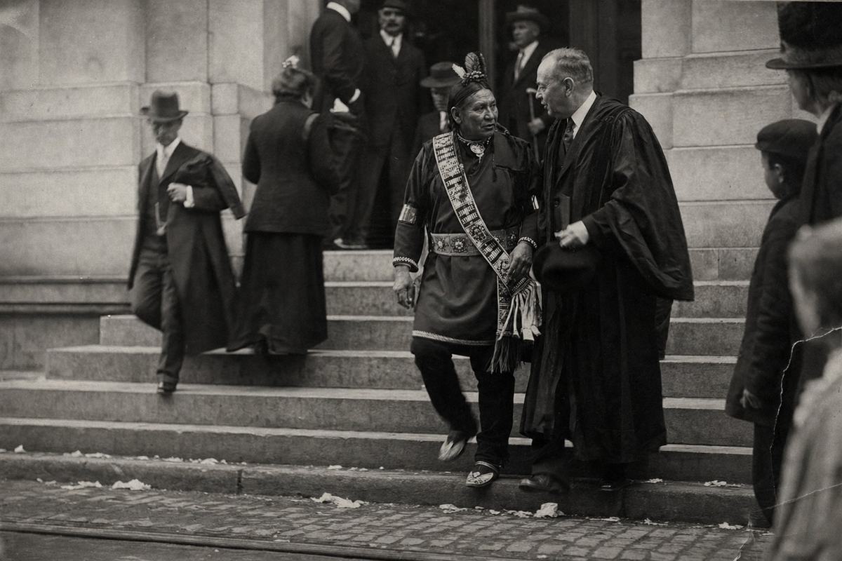 A silver gelatin print (c. 1909) capturing a candid moment at the inauguration of the Lenape Council Hall on Broad Street. The main subject is a sachem in a beaded sash stepping down the stone stairs and turning to speak with a robed alderman beside him; their faces are distinct, with different ages and builds. The street below shows uneven cobblestones, faint trolley tracks, and scattered paper scraps near the curb. Onlookers crowd the edge of the frame, some half-cut off; no readable signage. Tonal range is rich with deep blacks and soft midtones, slight edge silvering and a faint crease at the lower right; fiber-based paper with light selenium toning. View made with a 5x7 view camera from a low angle, slight motion blur in swinging coats, and shallow depth of field that softens the far doorway.
