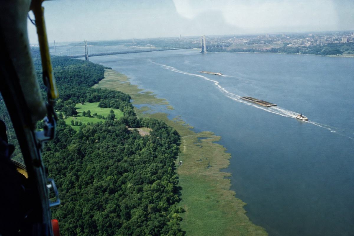 A 1968 aerial photograph on Kodachrome 64 color film showing the Trust Lands greenbelt at Inwood and preserved tidal flats, shot obliquely from a helicopter with the door open. The dominant subject is the sinuous line of marsh and upland trees at the northern tip of the island, placed in the lower left third; the Hudson estuary fills the frame to the right with two barges and a tug leaving white wakes. Colors display classic Kodachrome saturation with a slight cyan shift in the sky and deep, clean greens in the park canopy; fine, tight grain and a hint of vignetting at the corners. The George Washington Bridge is a small, hazy presence far in the background; no readable text anywhere. Captured with a Nikon F and a 105mm lens at 1/500 sec, minor motion blur from rotor vibration and a touch of window glare along the top edge.