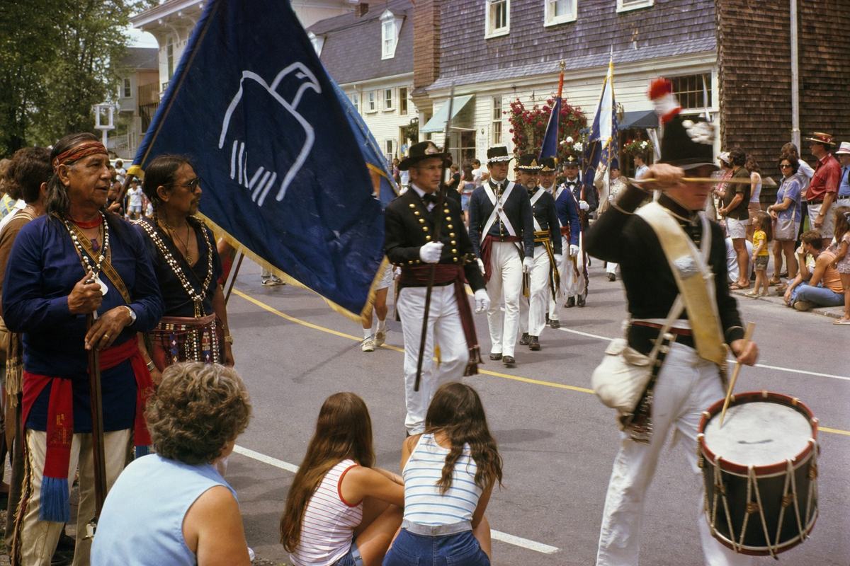 Candid street photograph on Kodachrome 64 color slide, 35mm SLR with 50mm lens, July 1976; joint Pequot–Connecticut commemorative procession moving along a gently curving village street near Mystic; Pequot color guard at left carrying a dark blue treaty flag with a simple white motif, Connecticut Guard detachment mid-frame; teenagers and elders in mixed summer dress watching from the curb; shingled storefronts and clapboard houses in the background with weathered paint and hanging flower baskets; breeze lifting small hand-held flags; warm color cast with saturated reds and clean blues, fine grain; slight motion blur in a drummer’s raised arm; no legible text anywhere.