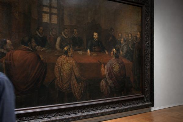 Color photograph taken in 1976 inside a quiet museum gallery of a seventeenth-century oil painting depicting the Lima cabildo in session with Diego de Almagro the Younger at the far end of a long table and two Inca envoys in rich mantles with sun-disc pins seated opposite. Shot on Kodak Ektachrome 64 with a Nikon F2 and 50mm lens at f/2.8; slight cool cast, fine grain, and gentle vignetting at the corners. Asymmetric composition from just left of center, with the dark wood frame cutting into the right edge and a faint reflection of a passerby’s shoulder in the varnish near the lower left. The canvas shows aged varnish, soft craquelure, and mellowed reds; gallery lights create small specular highlights without glare. Neutral wall, a stretch of polished floor at the bottom edge, no labels or signage in view.