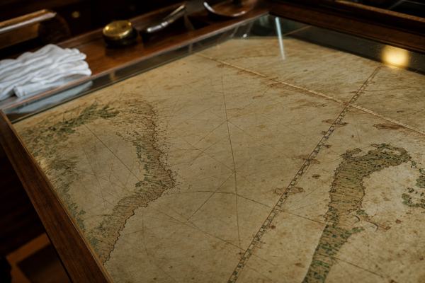1974 color photograph on Kodak Ektachrome 64, taken in a quiet museum reading room in Seville; oblique close-up of a 16th-century Iberian nautical chart laid under glass, showing coastlines of Brazil and West Africa with rhumb lines and a faint meridian; no visible lettering or labels; shallow depth of field; 50mm lens on a Nikon F2; raking tungsten light emphasizes parchment texture, stitch repairs, wormholes, and foxing; slight reflections on the glass; background hints of wood display tables, linen gloves, and a brass lens cap out of focus; asymmetrical framing with the meridian cutting diagonally across the frame.