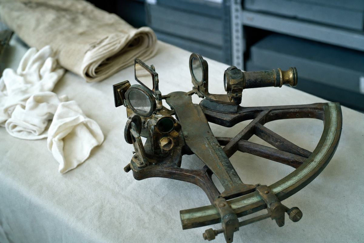 Color photograph of an 18th-century wooden and brass octant on a muslin-covered table, with conservation gloves nearby and no visible lettering.