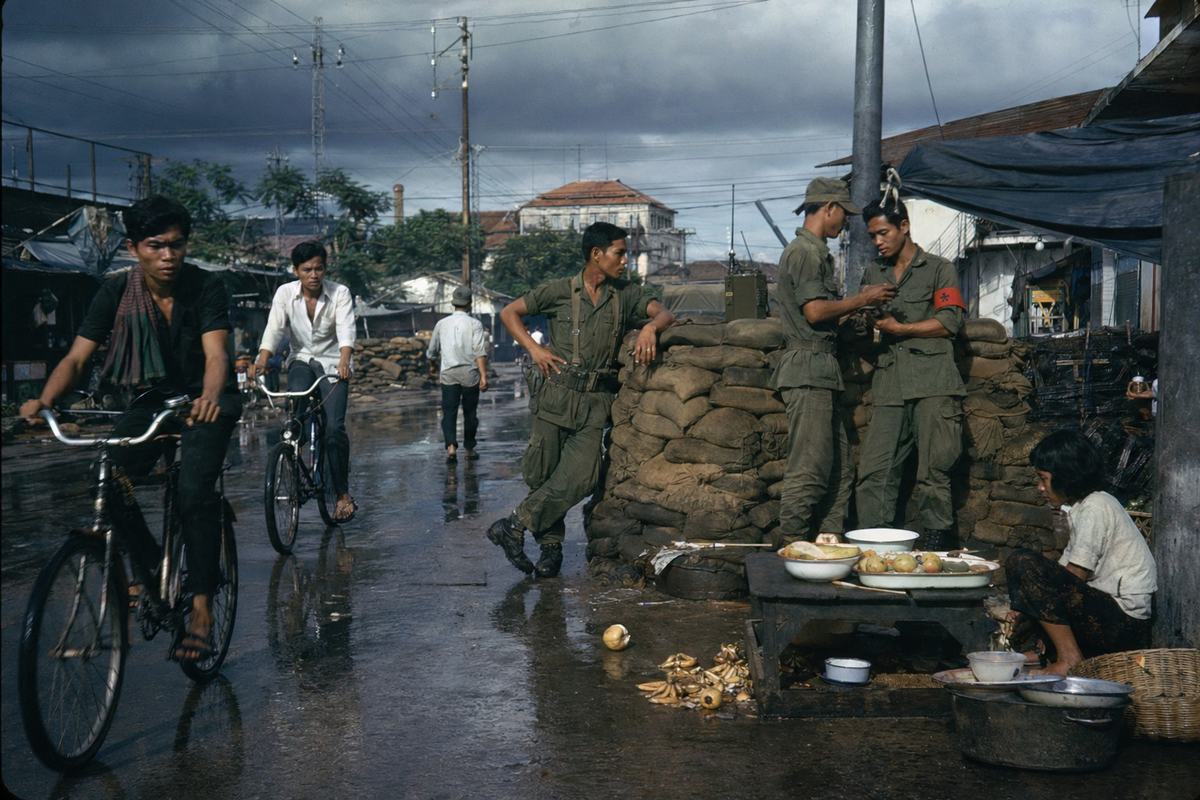 1973 color slide photograph on Kodachrome 64, saturated reds and warm skin tones with slight cyan in shadows; 50mm lens on a Leica M4, eye-level; candid Phnom Penh street scene near a sandbagged checkpoint under heavy monsoon clouds; two Cambodian civilians on bicycles weave past a guard in olive fatigues leaning on a sandbag wall, a liaison pair with colored armbands (no readable text) conferring by a radio set; puddles reflect tangled power lines, a street vendor’s stall with enamel bowls, scattered fruit peels on wet pavement; off-center framing with uneven exposure where sun breaks through cloud; distinct faces of different ages and builds, natural posture; no signage or lettering legible.
