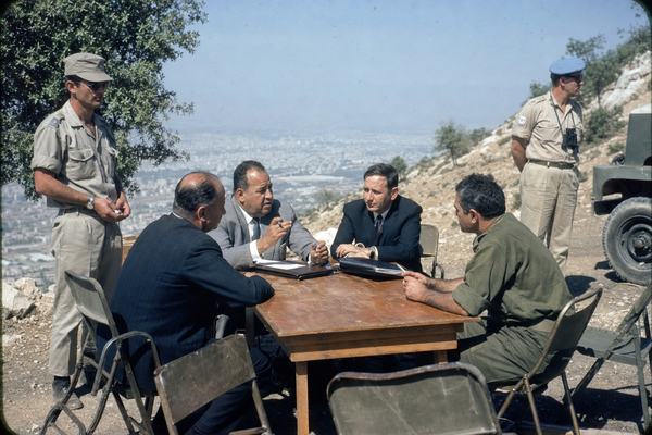 Color photograph on 1968 Kodachrome 64 slide film, taken with a Nikon F and 50mm lens at f/8 on the slopes of Mount Qasioun above Damascus; candid, asymmetric composition with a modest wooden table slightly off-center where two Syrian and two Israeli delegates sit, flanked by two UN observers in khaki; delegates mid-gesture with pens and closed folders, no visible lettering; natural midday light with city haze below, scrubby hillside and scuffed camp chairs; fine 1960s film grain, slight cyan cast, minor corner vignetting; a gust lifts a jacket cuff, a UN jeep edge intrudes at frame with no readable markings; realistic, varied faces and builds; no text anywhere in frame.