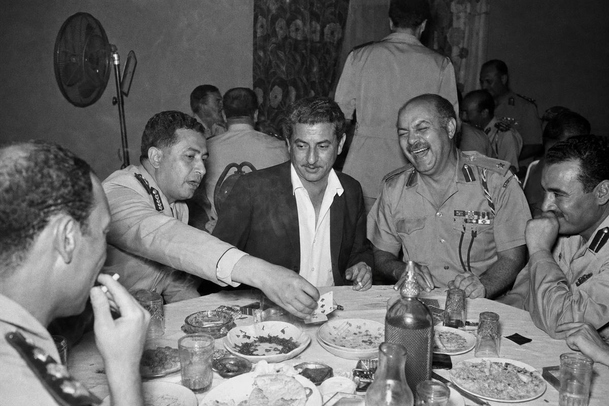 Black-and-white indoor photograph, 1964, taken on Kodak Tri-X 400 with a Leica M3 and 50mm lens, using a single flashbulb bounced off a yellowed ceiling at a Damascus officers’ reception; candid, off-center composition of Kamel Amin Thaabet seated at a crowded round table with Syrian officers in dress shirts and open-collar uniforms; cigarette smoke haze, glass ashtrays, half-eaten mezze plates, a sweating soda bottle; uneven flash falloff, pronounced grain, slight edge blur; one colonel mid-laugh, another reaching for a playing card; wall fan and patterned curtain in background; medals and epaulets visible but with no readable insignia; natural skin texture and varied ages; no text in frame.