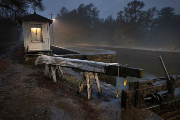 Digital press photograph taken pre-dawn at Augusta Canal Lock 3, Georgia; tripod-mounted long exposure around 1–2 seconds at ISO 1600 using a 24mm lens on a full-frame camera. The keeper’s hut sits left midground with a single warm lamp glowing through a rain-streaked window; wet granite lock walls glisten; a large steel pintle rests unevenly on two battered sawhorses under a partly transparent plastic tarp with frayed cords; small yellow audit tags flutter from two ratchets near the open south gate mechanism. Light river mist drifts over still, dark water; the muddy towpath in the foreground shows fresh boot prints, scattered pine needles, and a damp glove near a puddle. Background pines and a dim utility pole fade into haze; slight blur in treetops from wind. Asymmetrical framing with the disassembled gate cutting in at lower right; no centered people, only implied presence (a dented thermos on the stoop). No text, signage, or watermarks visible.
