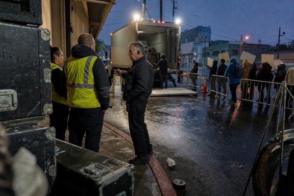 Digital press photograph captured on a full-frame mirrorless camera with a 28mm lens at f/2.8, ISO 6400, handheld; natural high-ISO noise in the shadows and slight motion blur on a passerby. Asymmetrical framing from just behind a stack of scuffed black road cases at the edge of a rain-slick loading apron. Scene: Bayview Civic Auditorium loading dock at Gate B on Toland Street at dusk; metal barricades and orange cones define a bent service corridor around a curb. Two Municipal Assembly Marshals in yellow safety vests confer with a venue supervisor wearing a radio earpiece beneath bright LED work lights. A small, disciplined protest line of 6–8 people stands back at a clearly measured distance from the service threshold; placards angled so no text is visible. One box truck with liftgate down is parked, puddles reflect the work lights, a crushed paper cup and a length of gaff tape lie near the curb. Background warehouse facades and utility poles soften into blue-hour haze. No visible text or logos anywhere in frame.