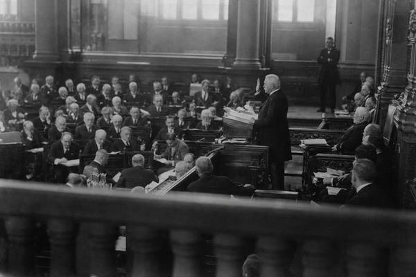 Archival black-and-white photograph, silver gelatin print from a 9x12 cm glass plate negative, Reichstag chamber, Berlin, May 1925; asymmetric composition from a side gallery balcony with a foreground railing slightly out of focus; Paul von Hindenburg in a dark civilian frock coat at a lectern, right hand raised mid‑oath; Gustav Stresemann visible two rows behind and to the left among dignitaries, head slightly bowed; delegates of varied ages and builds in formal suits, some turning pages; natural daylight spilling from high clerestory windows, dust motes visible; moderate grain, slight hand motion blur, soft falloff at corners; taken on a folding plate camera with a 135mm lens; incidental details include an usher leaning against a pillar and a stack of empty glass carafes near the aisle; no lettering or signage anywhere in frame.