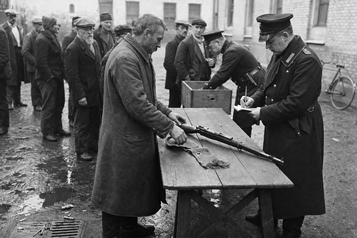 Archival black-and-white photograph, silver gelatin print from a 13x18 cm glass plate negative, barracks courtyard in Berlin, winter 1926; off-center view of a wooden trestle table where an ex-militia man in a worn overcoat places a bolt-action rifle and a frayed canvas armband; a uniformed police officer with a notebook and pencil records the handover while another officer checks a plain wooden crate; men queue loosely behind, faces distinct, varied ages and postures, caps and scarves; wet cobblestones with shallow puddles, a bicycle against a wall, cigarette butts near a drain; flat overcast light, uneven exposure toward the bright courtyard; fine grain, shallow depth of field around the hands and rifle; lens approximately 90mm on a plate camera; no text or lettering visible.