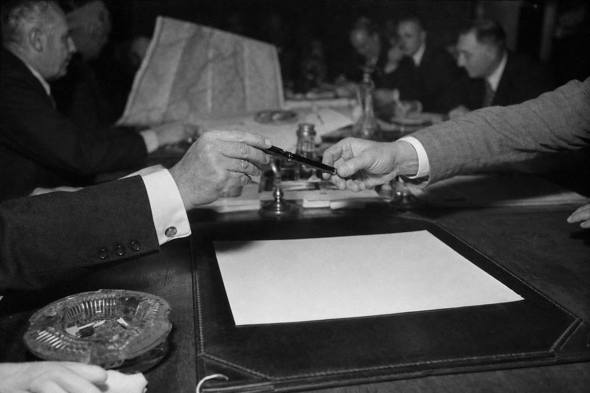 Black-and-white press photograph on 35mm nitrate film, Mainz, summer 1938; close, asymmetric framing on two distinct hands—one in a dark sleeve with cufflinks, the other in a lighter suit—meeting above a blank cream sheet placed on a leather blotter, fountain pen being passed; background out of focus shows a long table with inkwells, folded Rhine maps with no legible markings, and blurred faces of French and German delegates; single flashbulb produces hard shadows and slight glare on the paper; high contrast, visible grain typical of 1930s emulsions, slight vignetting; shot on a 50mm rangefinder; an ashtray with spent matches and a waiter’s white glove appear at the edge of the frame; no text or lettering visible.