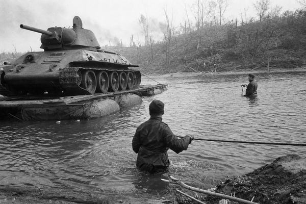 A historically accurate black-and-white press photograph (silver-gelatin fiber print) taken in May 1942 on Soviet Svema black-and-white film with medium grain, shot on a FED rangefinder camera with a 50mm Industar lens from a low muddy riverbank. Scene shows Red Army T-34 tank being guided across the Seversky Donets near Izium by sappers waist-deep in water, ropes taut, canvas pontoon segments bumping in the current. Asymmetric, off-center composition with the tank’s bow left of frame and two distinct sappers at differing distances; smoke haze and scattered branches in background, water ripples and splash droplets near the lens edge, slight motion blur on men’s hands. Overcast sky, uneven exposure along the waterline, no text or markings visible anywhere in the scene.