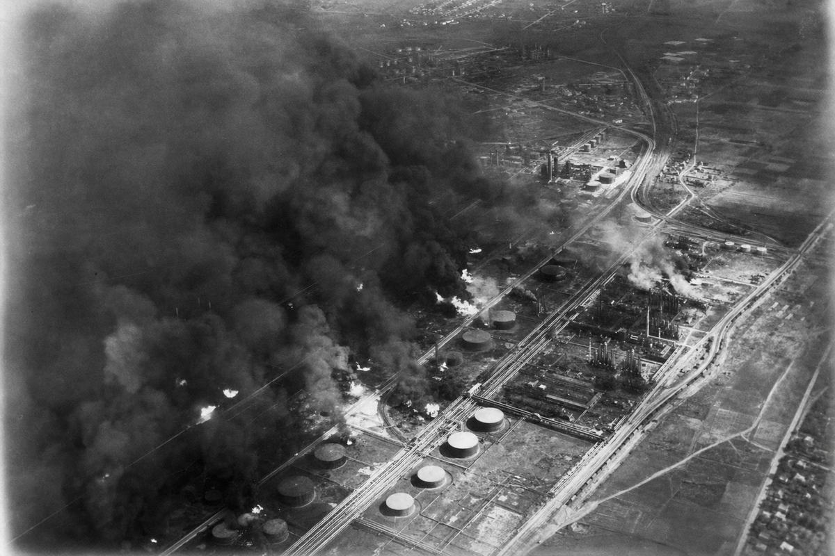 Oblique aerial reconnaissance black-and-white photograph (silver-gelatin contact print on glossy fiber paper) taken in July 1943 from an Allied reconnaissance aircraft using a K-20 aerial camera with a 152mm lens; fine-to-medium grain with slight edge blur from airframe vibration and mild vignetting. View shows the Ploiești refinery belt with multiple oil tank fires and thick smoke columns drifting across rail sidings and cracking units; scattered firefighting streams barely visible. Asymmetric framing with the densest smoke off-center, diagonal rail lines, and tank farms partly obscured by haze. High-altitude summer haze softens contrast; no frame numbers, fiducial marks, or any text visible in the image.
