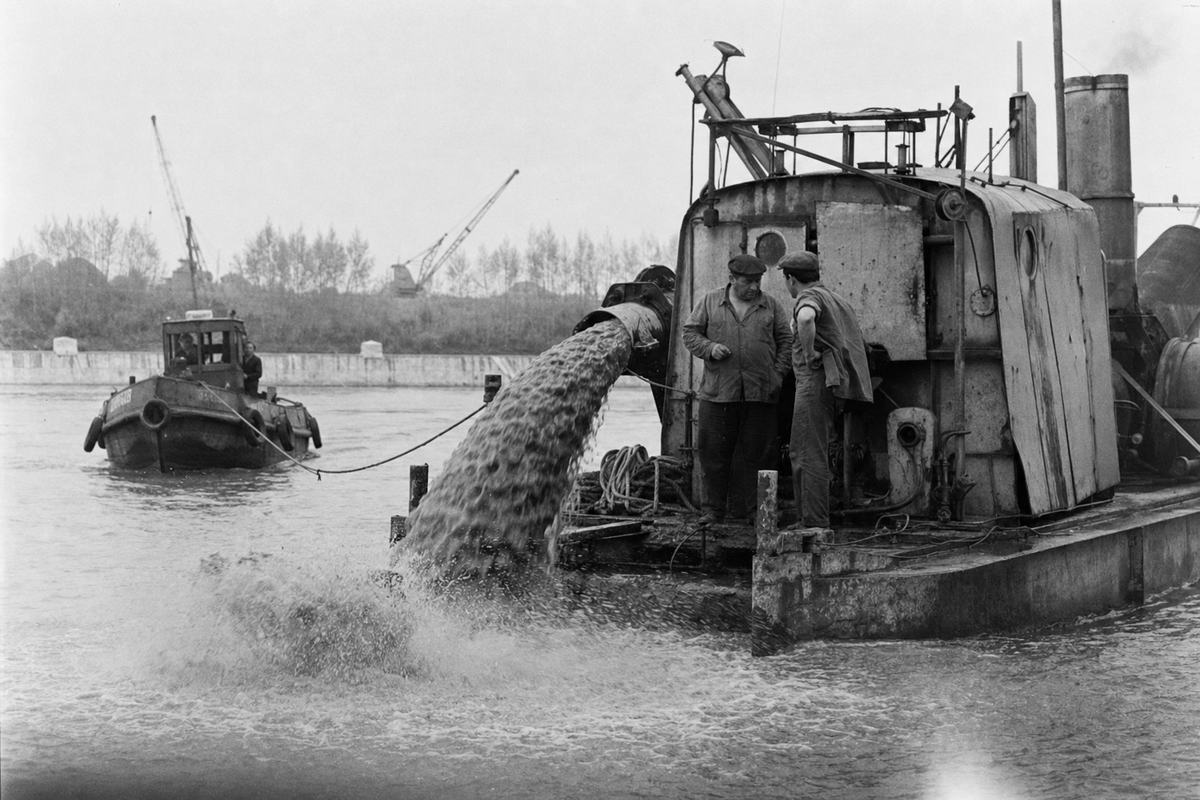 Late-1940s black-and-white industrial photograph (silver-gelatin fiber print) taken around 1949 on a Leica III or Zorki with a 90mm lens from a riverside embankment on the lower Danube near Brăila. Asymmetric composition: a suction dredger dominates right-of-frame, its discharge pipe arcing slurry back into the current, while a small tug strains at a towline left-of-frame; two deckhands of distinct build and age confer near a coil of wet rope. Misty overcast light, low contrast with crisp midtones; reed beds and a low quay with cranes fade into background; droplets and fine spray produce a soft flare along the bottom edge. No text, hull names, or markings legible anywhere in the image.