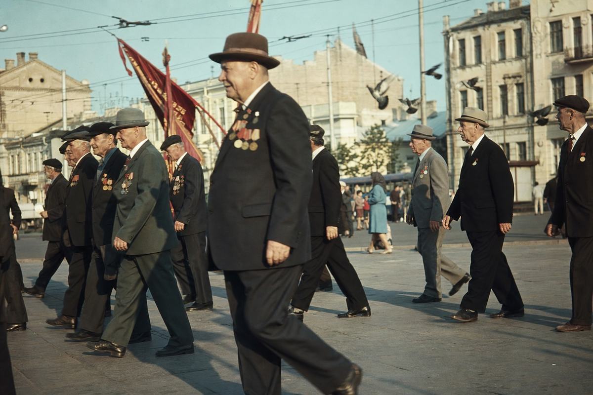 Early-1970s color slide photograph (ORWOchrome reversal film) with warm, slightly greenish cast and moderate grain, shot on a Zenit‑E with an 85mm lens from waist height at an oblique angle across Kharkiv’s central square. A file of aging veterans in dark suits and caps strides left to right across Ploshcha Dzerzhynskoho; one man mid-step in the foreground dominates, medals catching late-afternoon light. Distinct faces and builds among veterans; silk banners tilt backward in a light breeze with no legible text. Background shows trolleybus wires, pigeons fluttering up, and chalky apartment facades; shallow shadows across paving stones. Natural, candid posture; no modern signage and no lettering visible.