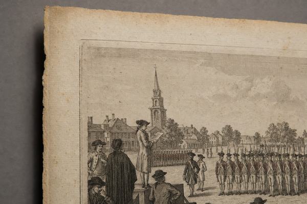 A museum studio object-study photograph shot on Kodachrome 64 (35mm) of an 18th-century copperplate engraving showing Boston Common with British redcoats mustering as a clerk reads a proclamation to townspeople. The print lies on a neutral archival gray backdrop; gentle raking light reveals paper foxing, deckled edges, and a pronounced plate mark. Composition is off-center, framing the upper-left quadrant of the print to exclude any cartouche or lettering; no text is visible. Fine film grain, slight warm cast, shallow depth of field from a 55mm micro lens; soft museum shadows at the edges of the sheet.