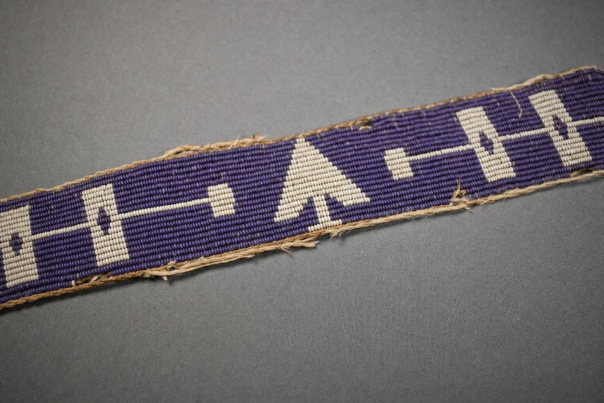 A 1970s copy-stand color photograph on Ektachrome 100 of a treaty wampum belt used in Prairie negotiations, woven in purple and white shell beads on sinew. Edges show light fraying and small areas of bead loss; surface has a soft matte sheen. The belt rests on a neutral gray muslin mount; photographed at a shallow oblique angle with raking light to reveal bead texture. Shot with a 60mm macro lens on a Nikon F2; fine grain, accurate color. No labels, tags, rulers, or text appear in frame; edges of the mount fall gently out of focus.