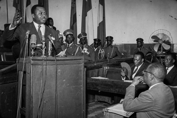 Black-and-white press photograph, silver gelatin print, July 1960, Elisabethville provincial parliament chamber during a heated session. Shot with a Leica M3 and 50mm Summicron on Kodak Tri-X 400; on-camera flashbulb creates harsh highlights and falloff, visible film grain and slight edge vignetting. Asymmetric composition from the left aisle: Moïse Tshombé mid-speech at a wooden lectern crowded with mid-century microphones, right hand raised, papers curling in his left hand; behind him hang plain flags and a row of uniformed officers in peaked caps. Suited officials sit on scuffed wooden benches in the foreground, one man fanning himself; parquet floor worn, dust motes caught in the flash, an oscillating fan with chipped blades in the background. No lettering, signage, or numbers visible anywhere.