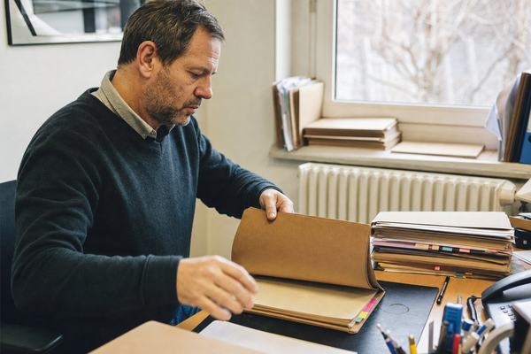 Digital press photograph, 2025, shot with a 35mm lens on a full-frame mirrorless camera. Natural winter window light. A middle-aged French civil servant sits at a simple office desk, three-quarter angle, off-center frame; he is mid-gesture opening a worn brown folder, not looking at the camera. Stacks of plain brown folders with colored index tabs are piled on the desk and the windowsill; a white radiator beneath the window; paperwork clutter, a pen cup, and a faint coffee ring on the blotter. Shallow depth of field isolates hands and folder; slight high-ISO noise in the shadows; a trace of motion blur in the fingertips. Asymmetric, candid composition. No text, logos, or legible labels anywhere.