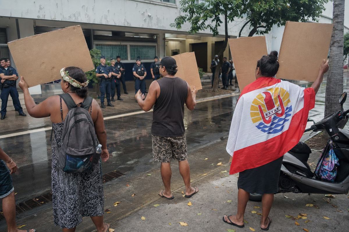 Petit rassemblement devant une antenne de l’État à Papeete; pancartes artisanales, drapeaux polynésiens, gendarmes au loin.