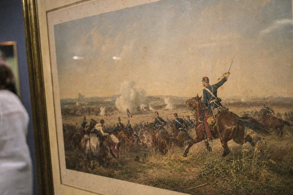 Archival reproduction photograph of a 19th-century chromolithograph depicting the cavalry charge near Morsbronn under a hazy summer sky; faded reds and blues of uniforms, drifting artillery smoke, trampled grass in foreground. The print shows yellowed paper edges, slight foxing, and a visible plate mark; frame edge intrudes at left. Shot obliquely to emphasize paper texture and minor glare from the glass; handheld, slight perspective skew. Background includes a sliver of a museum wall and the blurred shoulder of a passerby. 35mm Kodachrome 64, warm color cast, moderate grain, no text or labels visible, asymmetric composition.