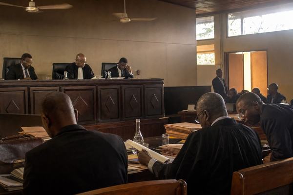Digital press photograph, late afternoon inside an arbitration courtroom in Abidjan; three arbitrators at a dark wooden bench under spinning ceiling fans; warm side light pouring through high windows; counsel tables cluttered with stacked manila files, water carafes, and scuffed briefcases; an aide leaning in to whisper while a lawyer flips a page; candid, off-center composition from aisle level; 35mm prime lens on a full-frame DSLR; shallow depth of field isolating the bench; ISO 3200 with fine luminance noise in the shadows; slight motion blur on a rotating fan; dust motes visible in the light beam; natural color; no visible text, signage, or logos anywhere.