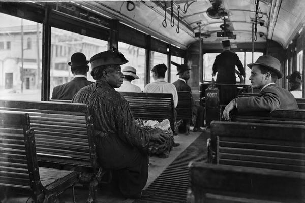 Silver gelatin print from dry-plate glass negative, circa 1898, oblique interior view from the rear platform of a New Orleans streetcar with Black and White passengers seated together; 8x10 view camera, 180mm rectilinear lens; mid-contrast tonal range, visible fiber-based baryta paper texture, slight silver mirroring at edges, minor corner wear; natural window light with uneven exposure and faint motion blur at the open windows; asymmetric composition centered on an older Black woman with a wicker basket on her knees while a younger White clerk across the aisle leans back with one forearm over the seat; scuffed wooden bench slats, metal fare box by the conductor, ceiling fans and leather hand straps; humid street glimpsed through windows; no legible signage or text anywhere; authentic period clothing and distinct faces with natural skin texture.