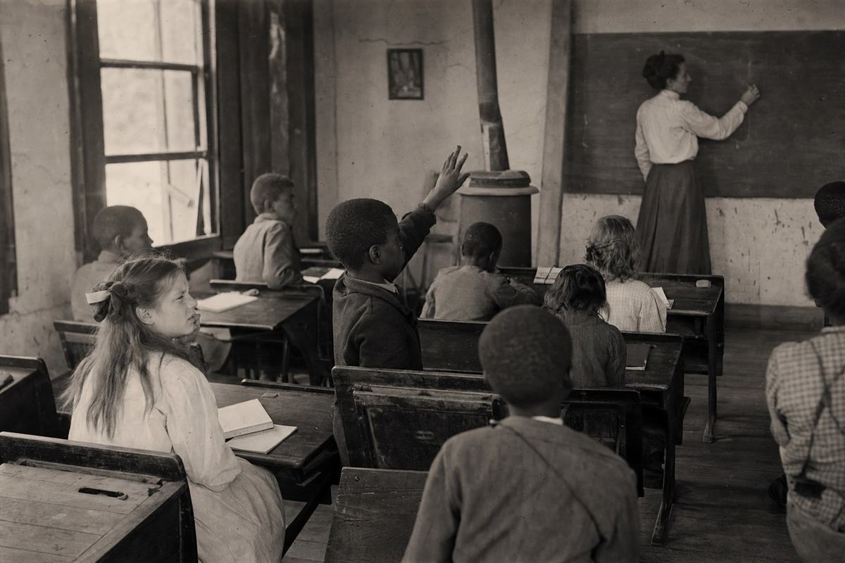 Silver gelatin classroom photograph, Louisiana public school, circa 1906; dry-plate negative contact printed on matte fiber paper; soft low-contrast tones with chalk-dust haze; 5x7 view camera, 150mm lens positioned in the back corner for a diagonal perspective; candid moment of an integrated class: a Black boy mid-gesture raising his hand while a White girl at the adjacent desk squints toward the teacher; desks nicked and ink-stained, open windows with warped frames, scuffed floorboards, small coal stove at rear; teacher (middle-aged woman) turned three-quarters writing faint marks on a slate board with no legible text; uneven exposure falloff at edges, slight emulsion scratches; no posed symmetry, distinct faces and ages.