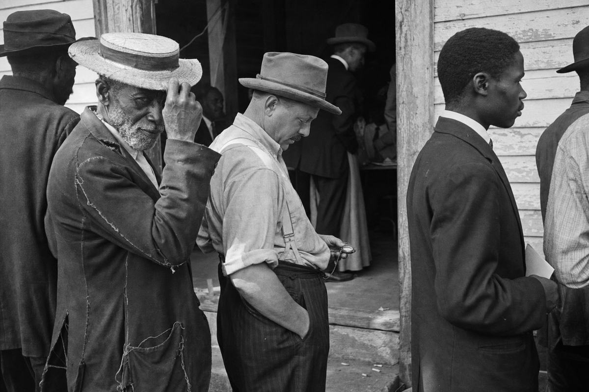 Gelatin silver press photograph, Southern polling place, 1912; nitrate negative printed on glossy fiber paper with pronounced grain and high contrast; handheld 4x5 press camera with 127mm lens at waist level; off-center view down a short queue of mixed-race voters: an elderly Black man in a frayed straw boater gripping the brim, a White mill worker with rolled sleeves checking a pocket watch, a young Black teacher holding a folded paper with printing turned inward so no text is visible; doorway in shade, poll officials glimpsed inside without signage; cracked sidewalk, cigarette butts near the step, peeling clapboard paint; slight motion blur on hands, uneven exposure across faces; candid, distinct expressions and builds.