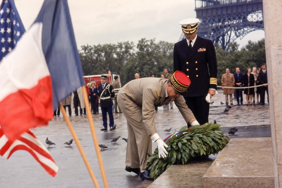 1977 color photograph on Kodachrome 64, 35mm SLR with 50mm lens; candid, off-center composition at the Champ-de-Mars during a Franco-American wreath-laying ceremony; dominant subject: a French officer in dress uniform bending to set a laurel wreath while an American naval attaché stands a step behind with head bowed; crossed national flags near the left edge with slight motion blur; medium grain, saturated reds and deep blues with warm Kodachrome bias; bright overcast sky, soft shadows; incidental background: lower lattice of the Eiffel Tower partly visible, a gendarme and a few onlookers behind a rope, pigeons on damp pavement; natural skin texture and varied physiques; no visible lettering on wreath ribbons, no signage or banners with text.
