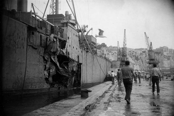 Black-and-white silver gelatin press photograph, Malta, June 1967; documentary style; the USS Liberty moored at Valletta with its port-side hull torn open above the waterline, twisted steel plating and scarred paint; sailors and Maltese dockworkers moving along a wet quay; cranes and weathered limestone fortifications in the background under soft Mediterranean haze; a gull crossing mid-frame; shot on 35mm Kodak Tri-X 400 with a Nikon F and 50mm lens; moderate grain, slight motion blur on figures, edge vignetting and uneven exposure; asymmetrical composition from shoulder height on the quay with the ship offset to the left third; no visible signage or lettering; archival print scanned with light surface scratches and dust.