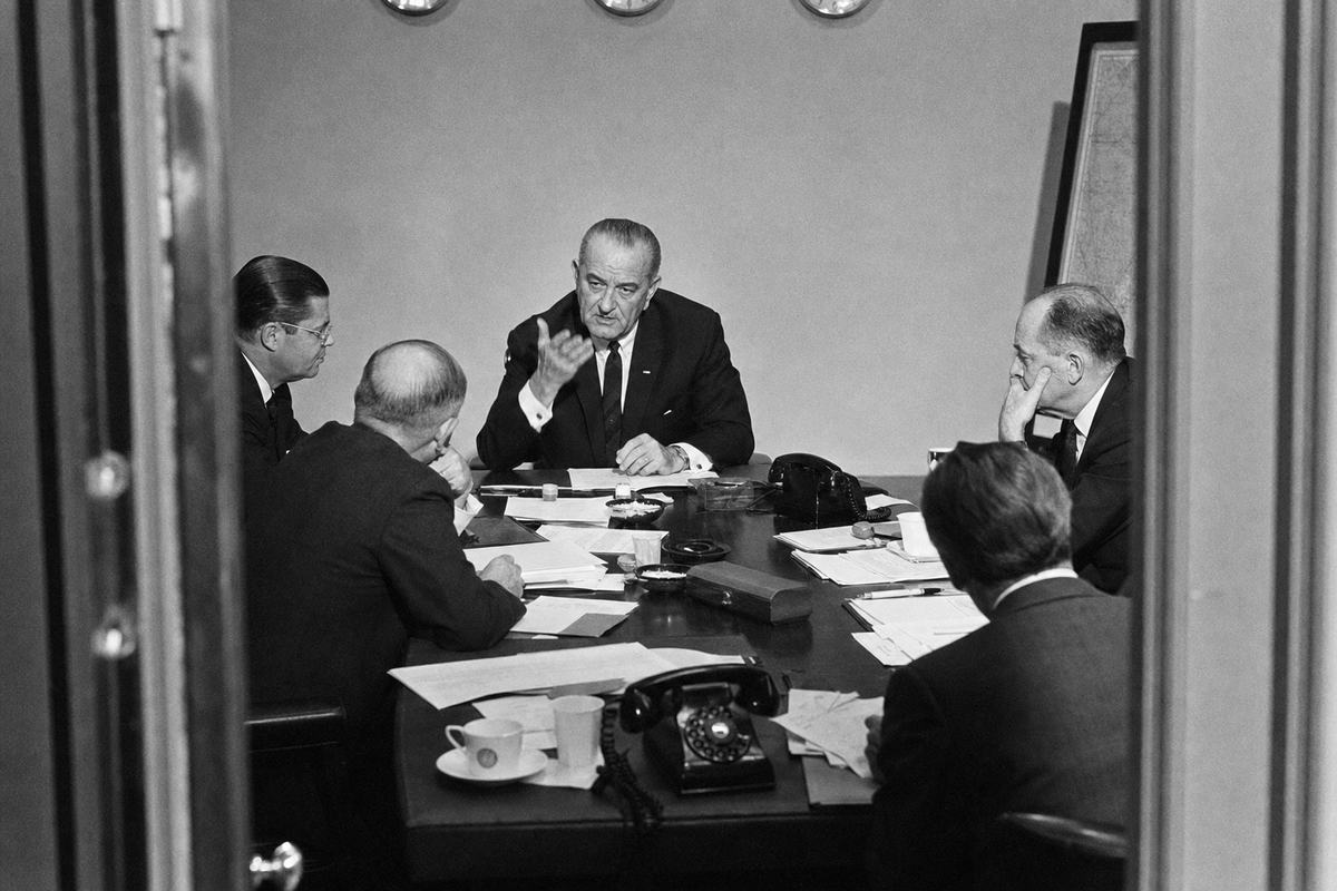 Black-and-white press photograph inside the White House Situation Room, June 1967; President Lyndon B. Johnson at the head of a narrow table with Robert McNamara, Dean Rusk, and Walt Rostow leaning over scattered papers; ashtrays, rotary telephones, and coffee cups visible; wall clocks half out of frame; a map board faintly visible with no legible markings; flashbulb illumination with hard shadows; shot on 35mm Kodak Tri-X 400 with a Nikon F and SB-6 flash, 35mm lens; pronounced grain, slight motion blur in a gesturing hand; taken obliquely from the doorway for an off-center composition; no visible signage or text.