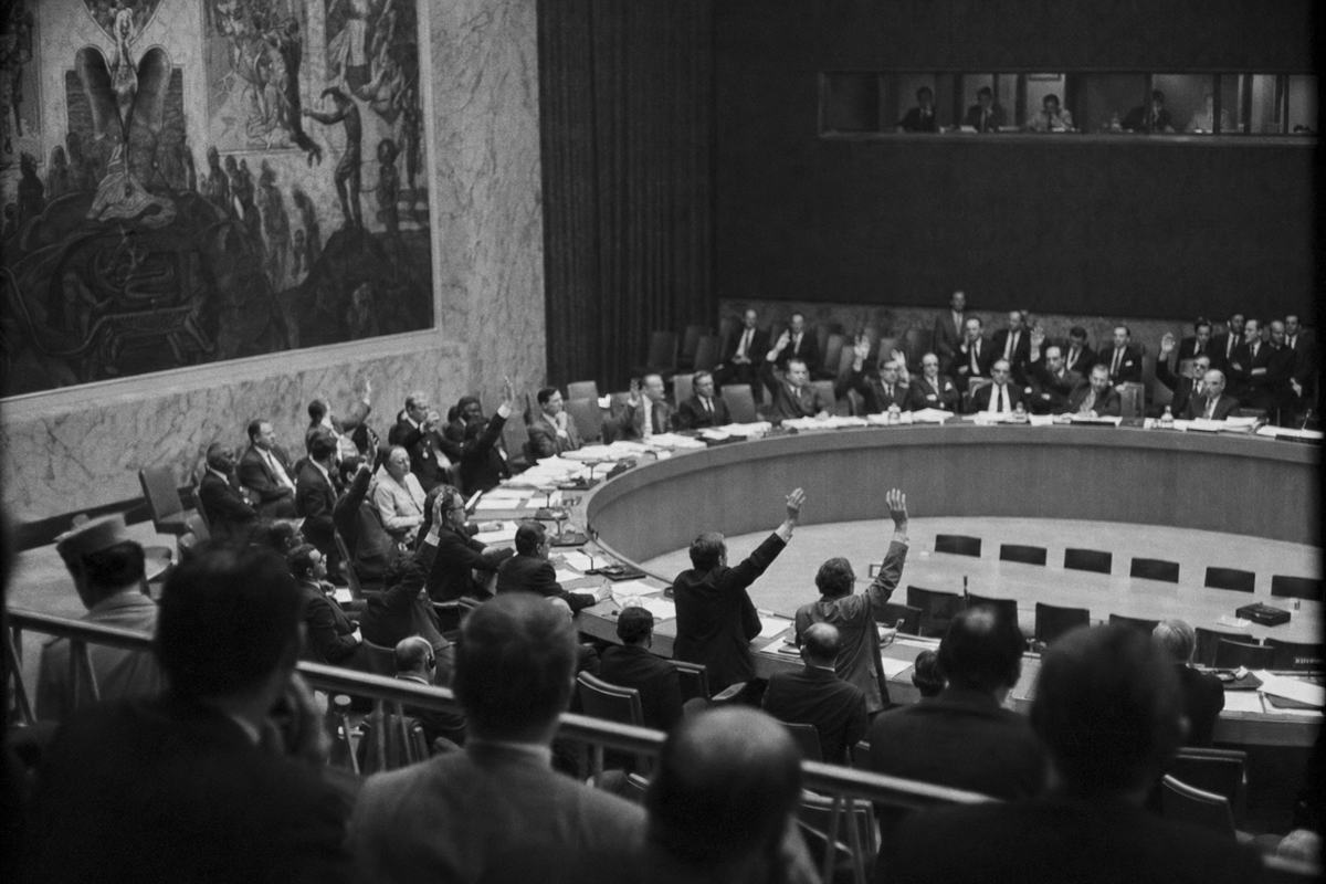 Black-and-white press photograph of the United Nations Security Council chamber, New York, 9 June 1967; delegates seen from the public gallery as hands are raised for a vote; interpreters behind glass and the chamber mural in the background; stacks of papers on the curved desk, none legible; tungsten lighting with gentle falloff; shot on 35mm Kodak Tri-X 400 with an 85mm lens; moderate grain, slight blur on moving hands, corner vignetting; diagonal composition with the horseshoe table sweeping across the frame; no readable nameplates or signage.