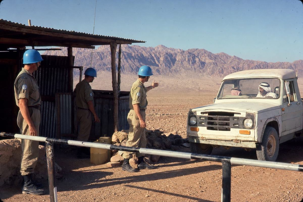 Color Kodachrome 64 slide, mid-1970s, Sinai desert checkpoint; a European patrol with blue helmets and desert fatigues stands beside a white-painted Land Rover without markings; a UN peacekeeper gestures to a Bedouin driver in a dusty pickup edging past a striped barrier; a corrugated-iron hut casts shade; jerrycans and a whip antenna nearby; late-afternoon light with long shadows and mild heat shimmer; saturated colors with a slight warm cast typical of Kodachrome; shot on a Leica M4 with a 50mm lens; fine grain, minor dust and scratch marks from archival slide scanning; candid, off-center composition with the vehicle entering from frame right; no signage or visible lettering.