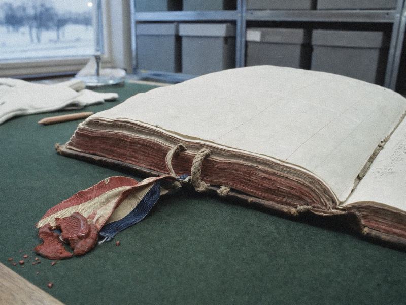 1970s color film photograph (Ektachrome 64), close-up of a 19th-century port register on an archive reading-room table; thick laid paper pages with chain lines, visible sewing on raised cords, red-dyed edges, a broken red wax seal on a tricolor ribbon; asymmetric composition with the ribbon and seal dropping into the lower-left foreground; incidental background details: green blotter, cotton handling gloves, a wooden pencil stub, out-of-focus grey archival boxes on metal shelving, cold winter window light; taken with a 50mm lens on a Nikon FE, shallow depth of field, slight blue cast and fine film grain; no visible text or labels anywhere.
