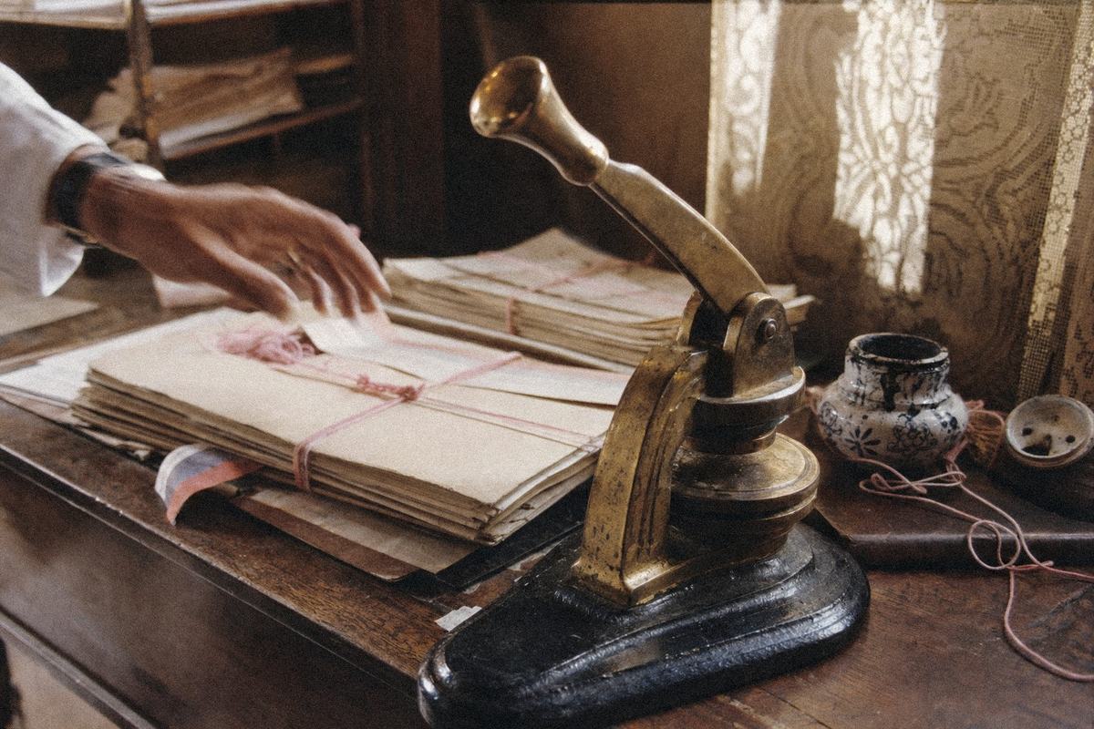 Presse à gaufrer en laiton sur un bureau, chemises cartonnées et ficelles roses; un ruban tricolore dépasse d’une liasse.