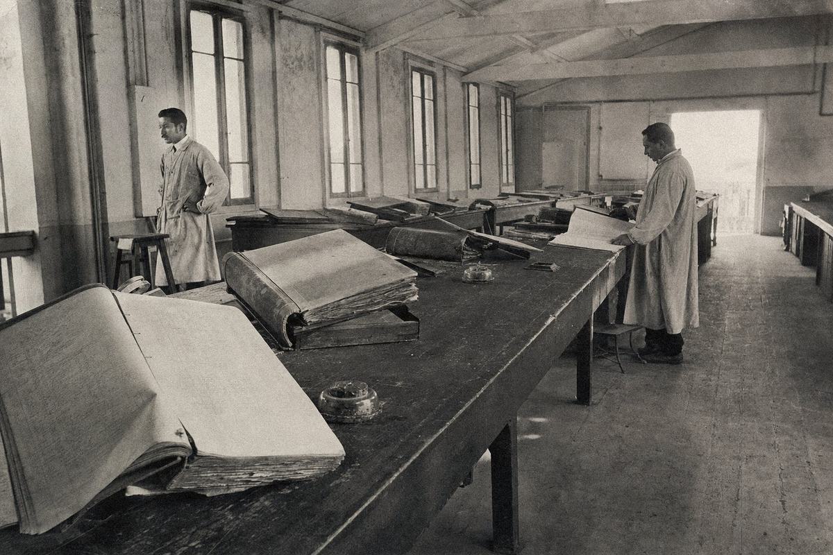Intérieur du lazaret d’Arenc à Marseille, pupitres, encriers et registres sanitaires alignés; deux agents en blouse claire.