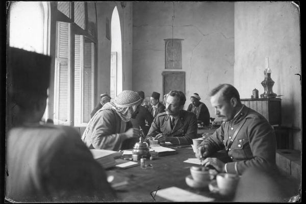 Archival gelatin silver print (dry plate), Damascus, 1918, inside a high-ceiling Ottoman-era reception hall with tall arched windows and wooden shutters; candid moment of a seated Arab delegate in keffiyeh leaning forward to address a French officer while a British officer scribbles notes; long tables cluttered with inkwells, folded papers, and teacups, no banners or visible lettering; soft window light from the left, slight motion blur in hands; shallow tonal range with milky highlights and deep midtones; shot on a 9x12 cm plate camera with an uncoated 150mm Tessar lens, slight edge falloff; soft film grain, minor emulsion scratches and corner wear; asymmetrical composition with a blurred foreground shoulder intruding; background shows plaster walls with hairline cracks and a brass oil lamp on a side table.