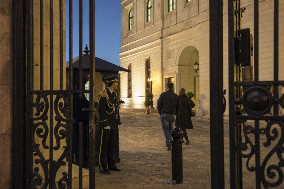 Digital press photograph, Aleppo, 2016, dusk; off-center view through an open iron gate toward the limestone facade of the Federal Council building; two civilians walk past a guard post while a uniformed ceremonial guard stands at ease, head turned toward arriving visitors; warm sodium streetlights mix with a cool evening sky; shallow depth of field with a 35mm lens on a full-frame DSLR, ISO 3200 noise visible in shadows; worn paving stones, a stray plastic bottle near a bollard; no plaques or lettering visible in frame.